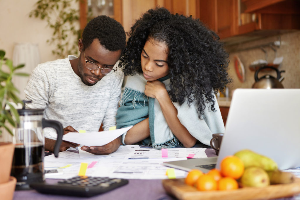 African male holding piece of paper while paying gas and electricity bills online on notebook pc. Young family calculating their expenses, planning domestic budget, sitting in kitchen interior