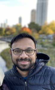 Sanket, smiling in glasses and a coat outdoors