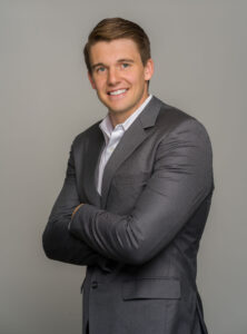 Tyler, standing with his arms crossed in a grey suit, smiling
